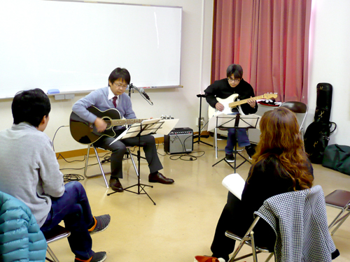 40代男性の生徒さんと講師のギター演奏とそれを聴く生徒さんの様子