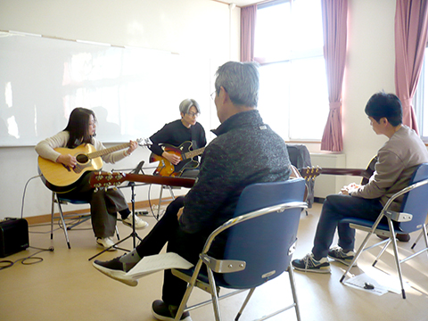 50代女性の生徒さんと講師のギター演奏とそれを聴く生徒さんたちの様子
