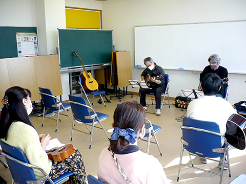60代男性の生徒さんと講師のギター演奏を聴く生徒さんたちの様子